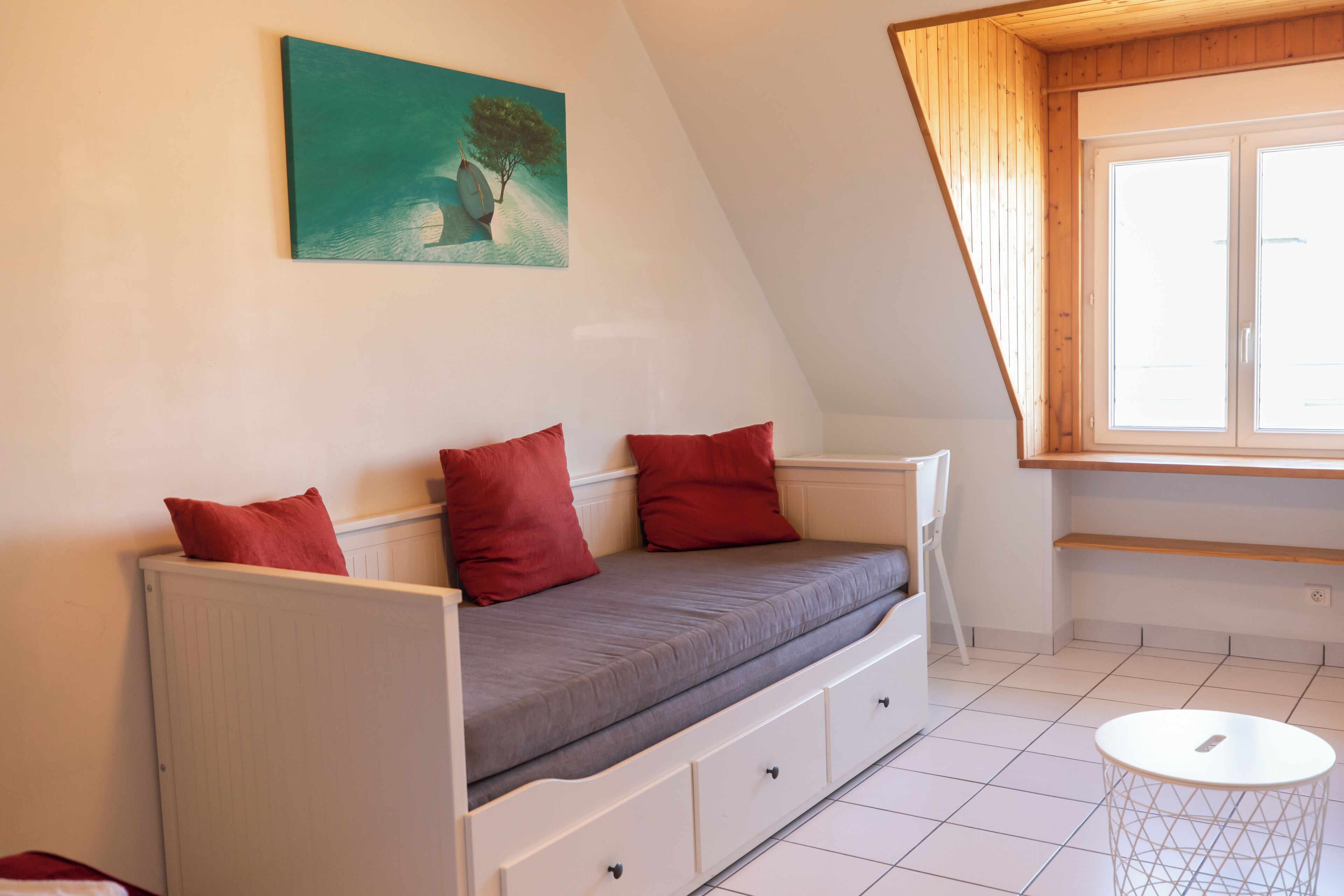 A small, cozy room with a sloped ceiling, a white radiator, a grey sofa with red cushions, a white round table, and a window with wooden shutters.