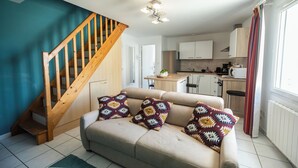 Un salon avec un canapé beige, des coussins à motifs, un escalier en bois et une cuisine à l’arrière-plan.