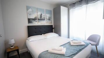 Una cama bien hecha con un edredón azul, una lámpara de noche y una silla junto a la ventana.