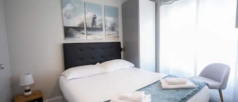 A neatly made bed with a blue blanket, a bedside lamp, and a chair by the window.