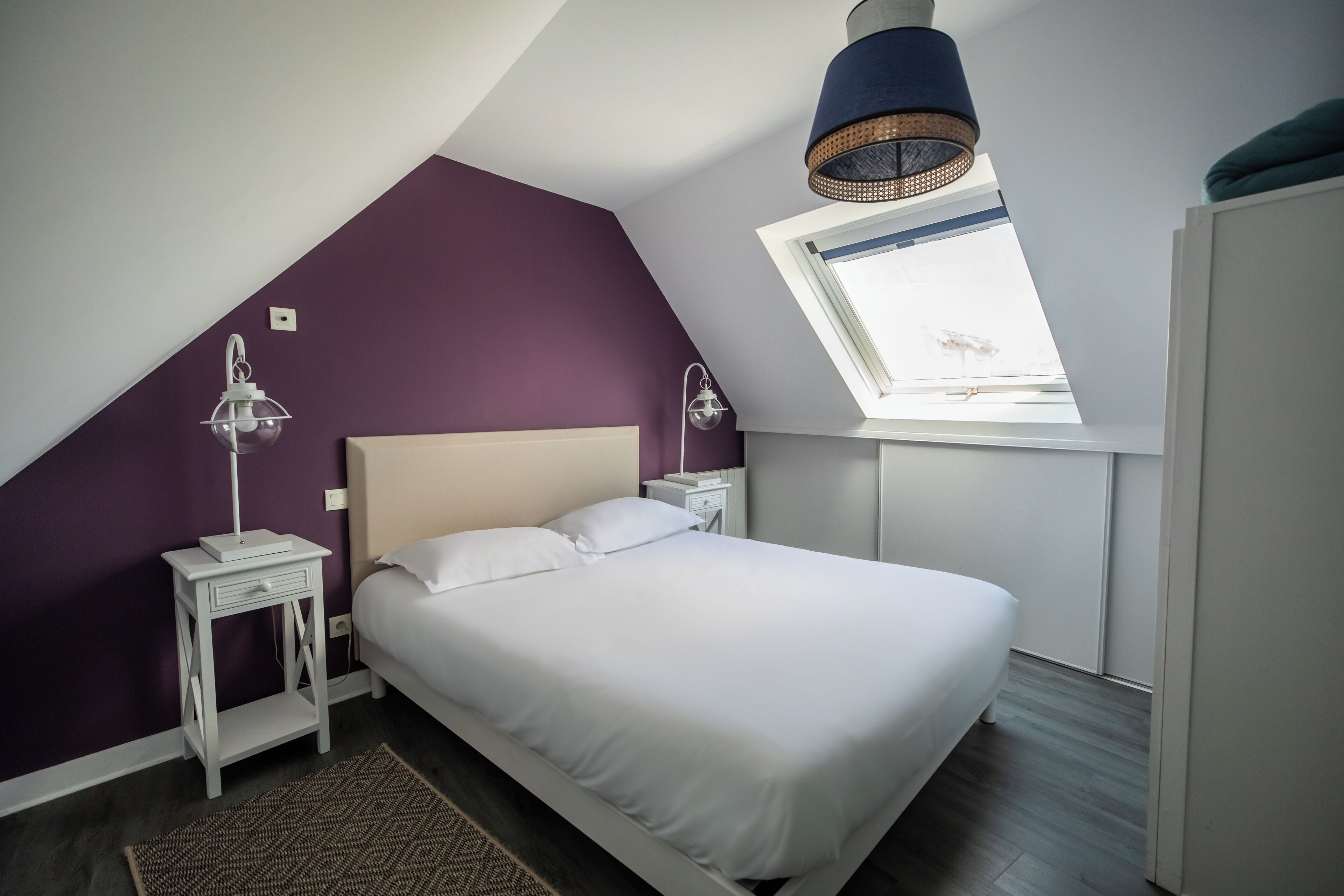 Un dormitorio con techo inclinado, una cama con ropa de cama blanca, dos mesitas de noche con lámparas y una ventana con vista al cielo.