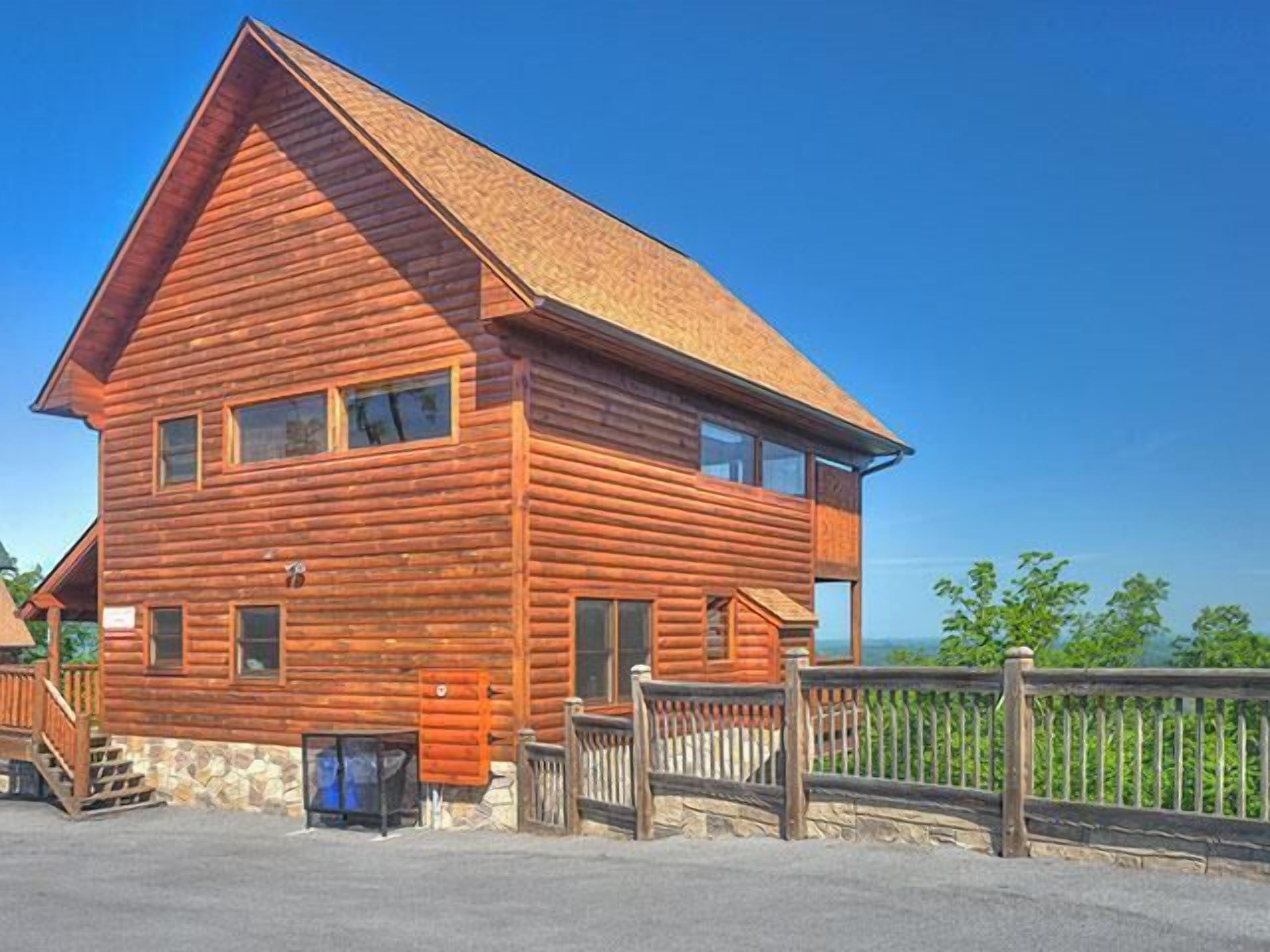 A two-story log cabin with a steep gabled roof, a stone foundation, and a wooden fence.