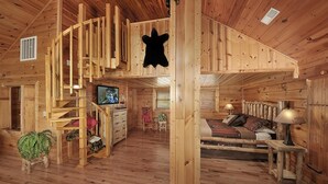Ein Holzhütten-Interieur mit Schlafzimmer im oberen Bereich, einer Treppe, einem Fernseher und einem Essbereich.