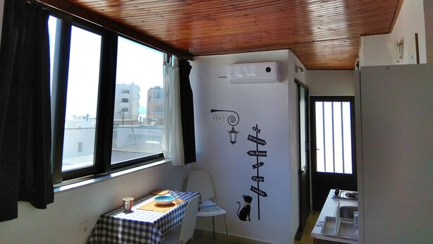 Une pièce avec un plafond en bois, une fenêtre avec des rideaux, un climatiseur fixé au mur, une table avec un bol et une tasse, et un mur orné d’une illustration de chat et de panneaux d’indication.