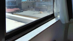 Une vue depuis une fenêtre, à travers un rideau, donnant sur un paysage urbain avec de nombreux bâtiments, dont un grand immeuble d’appartements et une construction plus petite avec un toit rouge.