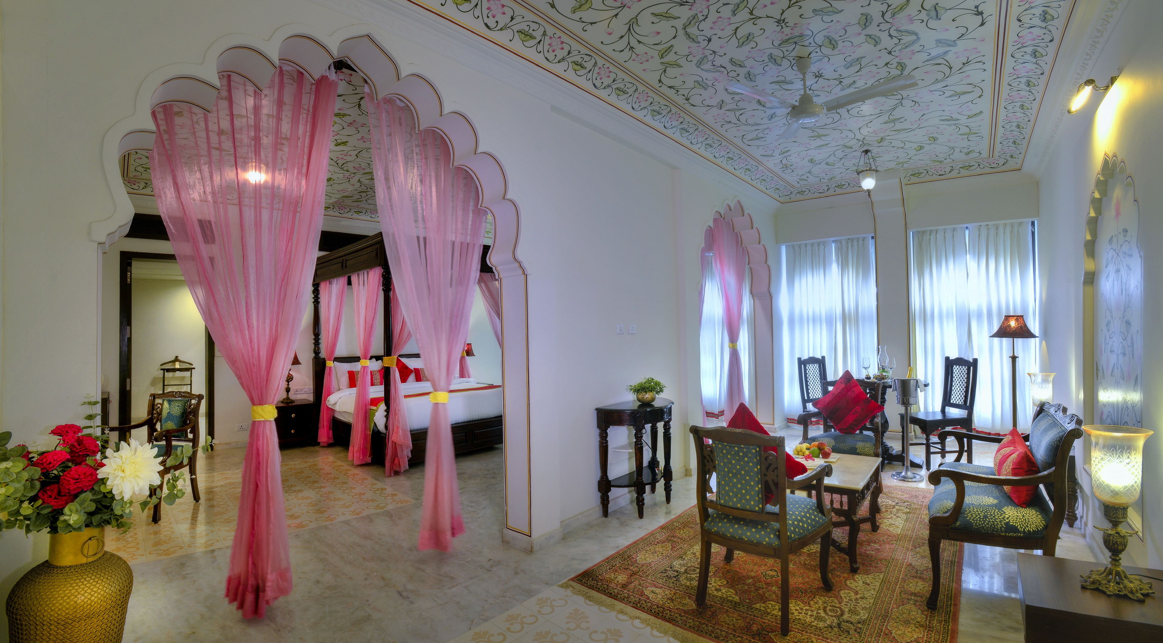 A room with a four-poster bed, a dining area with chairs and a table, and a decorative ceiling.