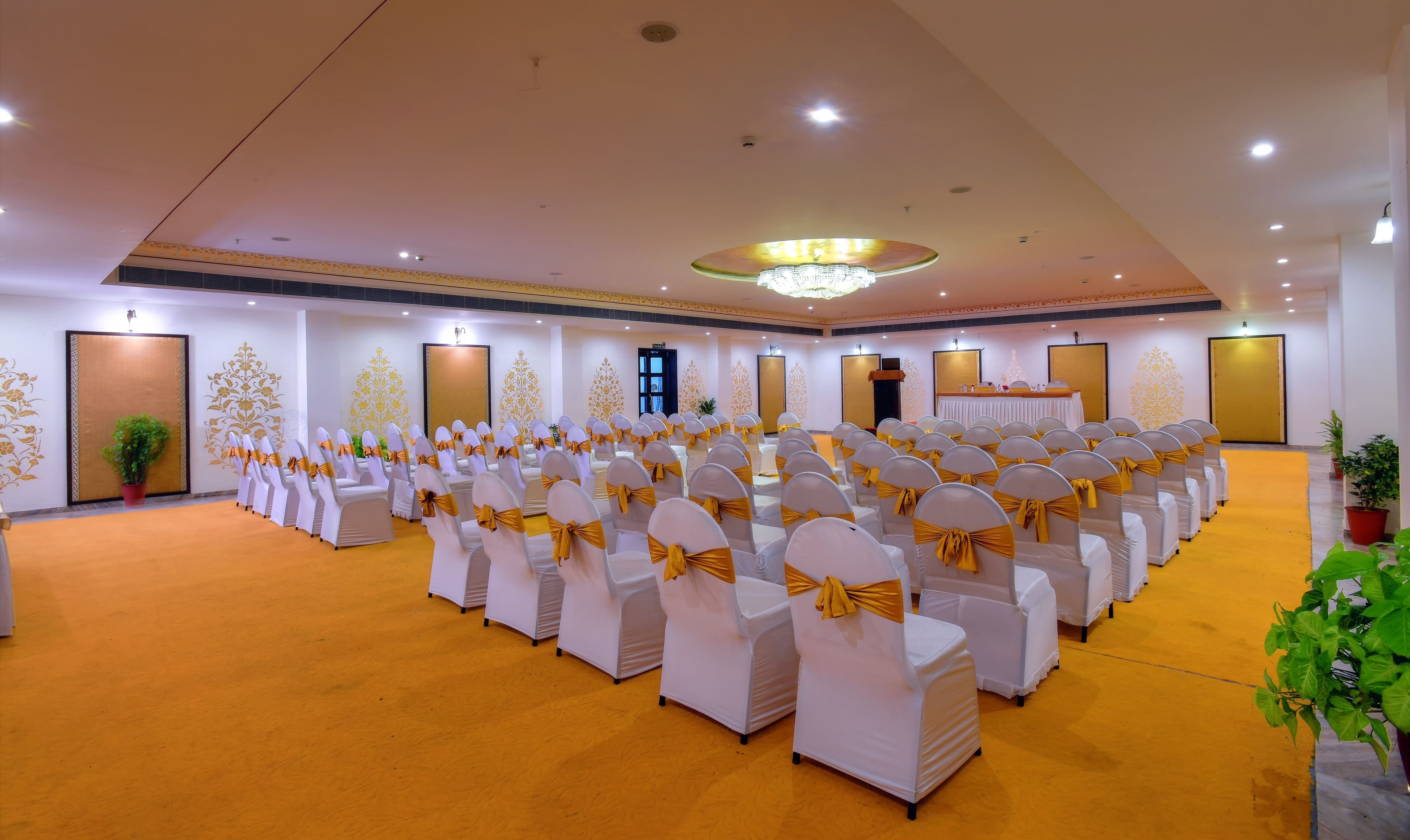 Une grande salle avec des rangées de chaises blanches ornées de rubans dorés, une scène avec une table et une moquette jaune.