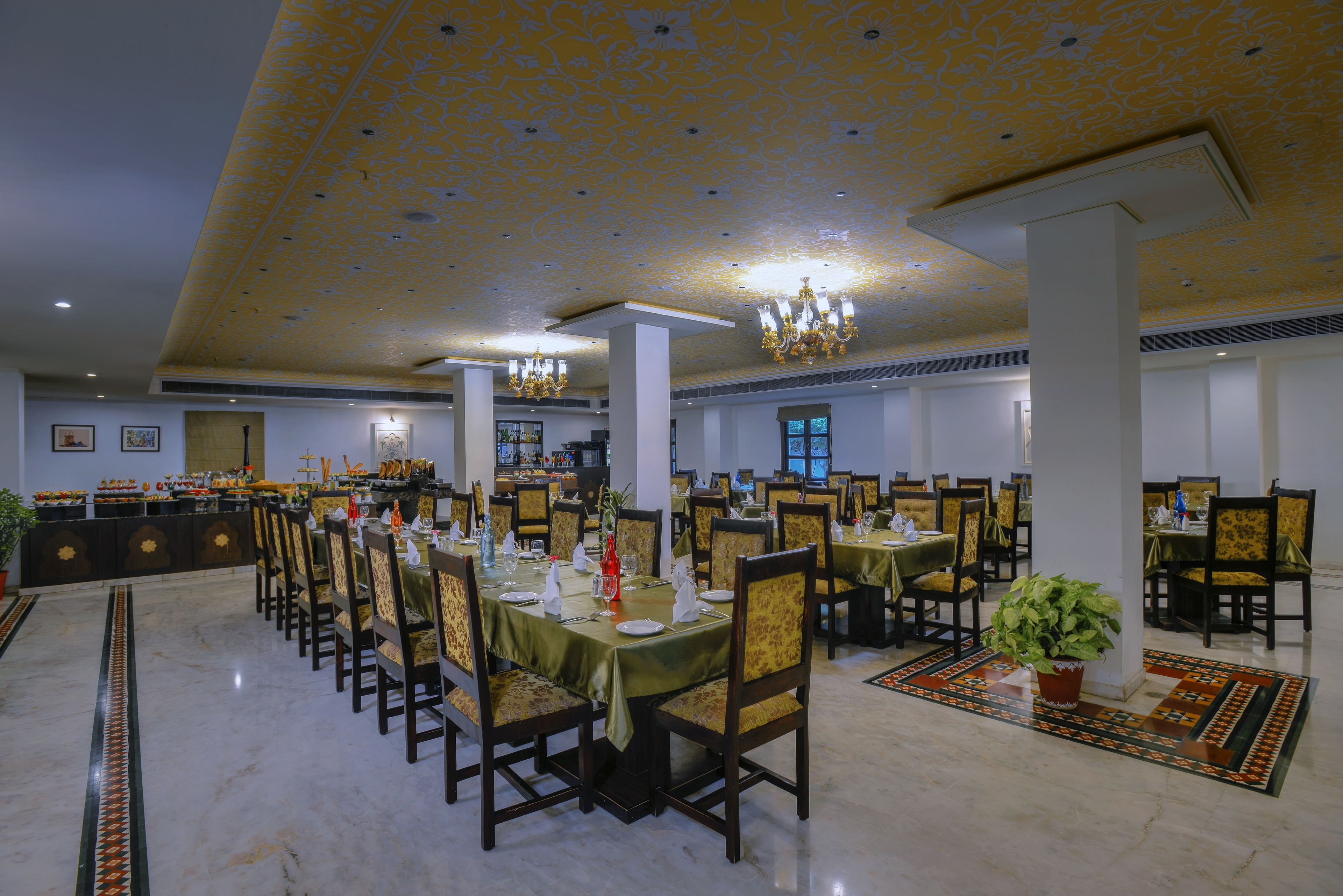 A spacious dining hall with multiple tables and chairs, a patterned ceiling, and chandeliers.