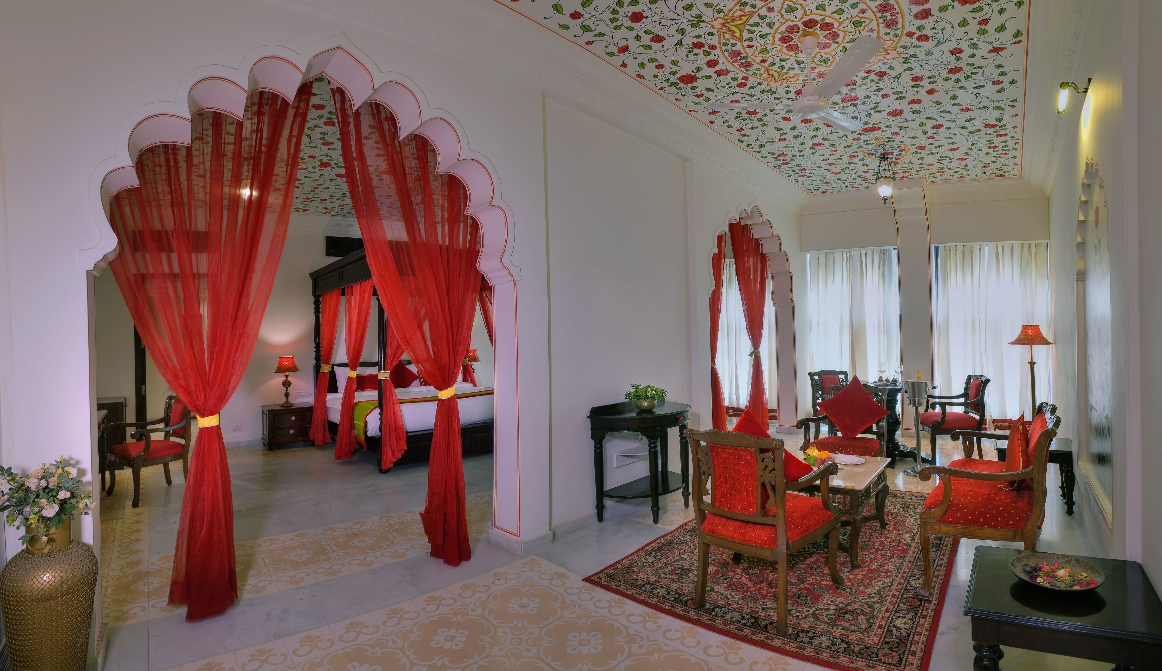 Una habitación con cama con dosel, cortinas rojas, un comedor con muebles de madera y un techo con motivos florales.
