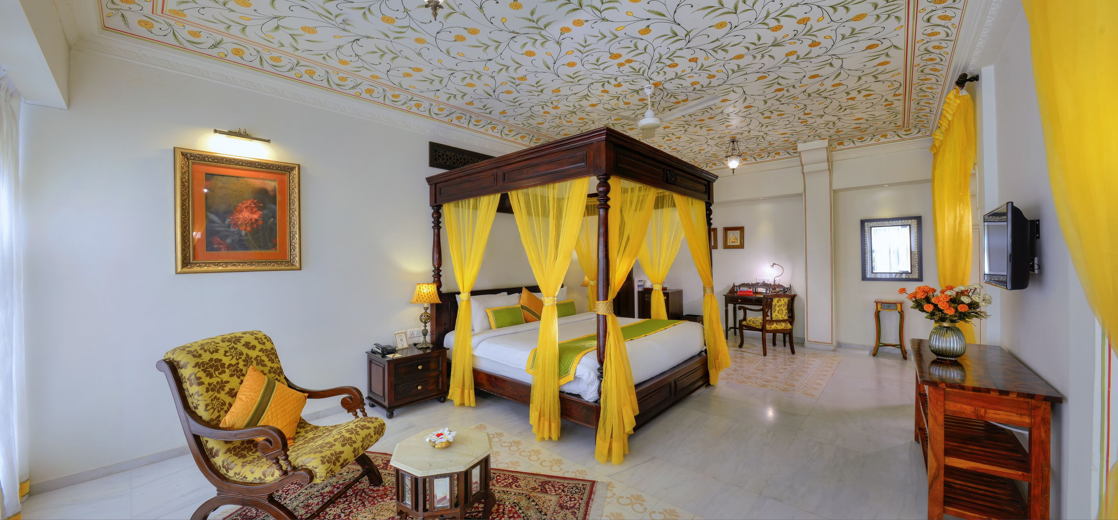 A four-poster bed with yellow curtains, a wooden chair, a small table with a vase, and a framed picture on the wall.