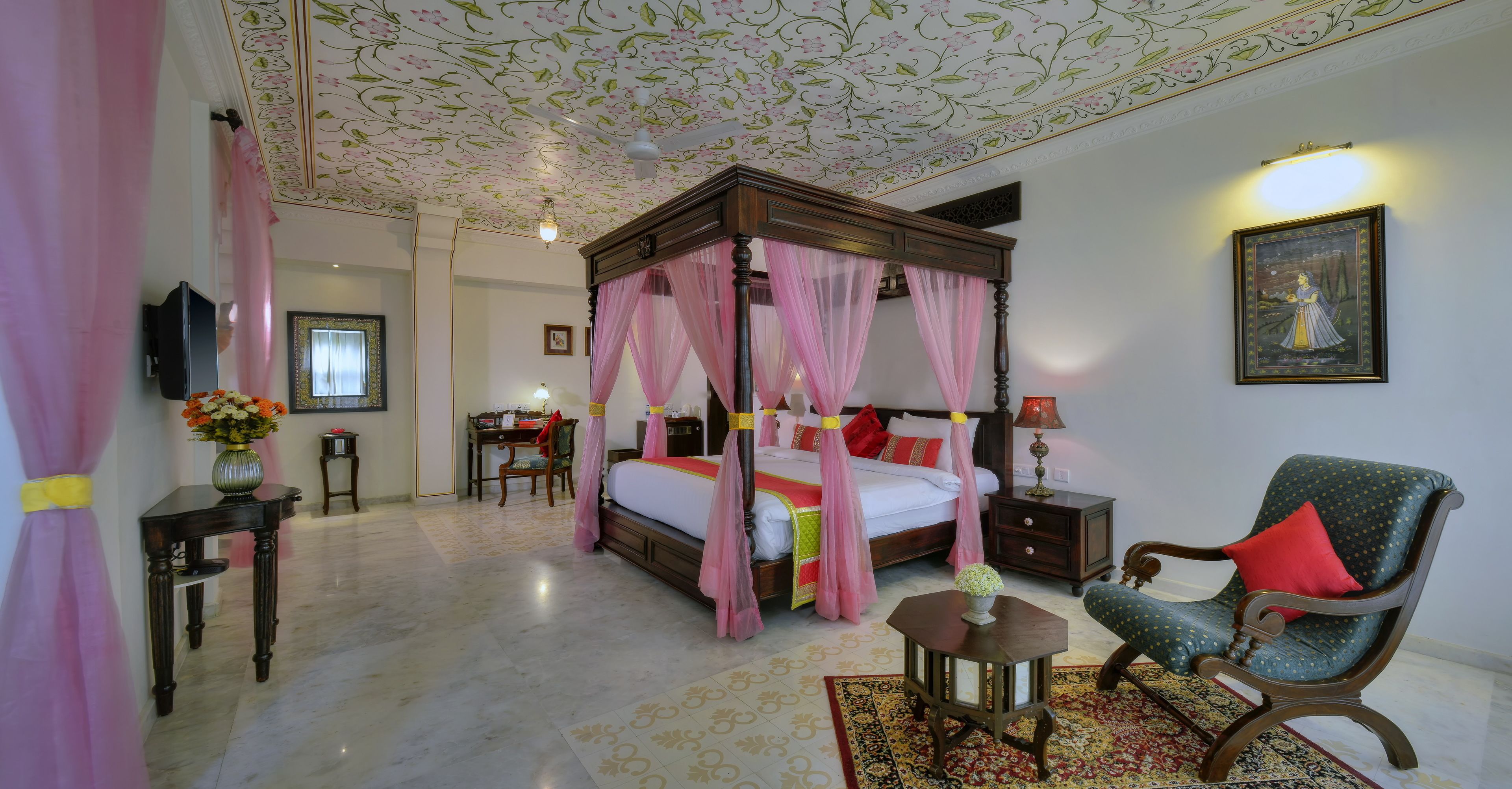 Un lit à baldaquin rose, une chaise en bois avec un coussin rouge, un bureau avec une lampe et un tableau encadré au mur.