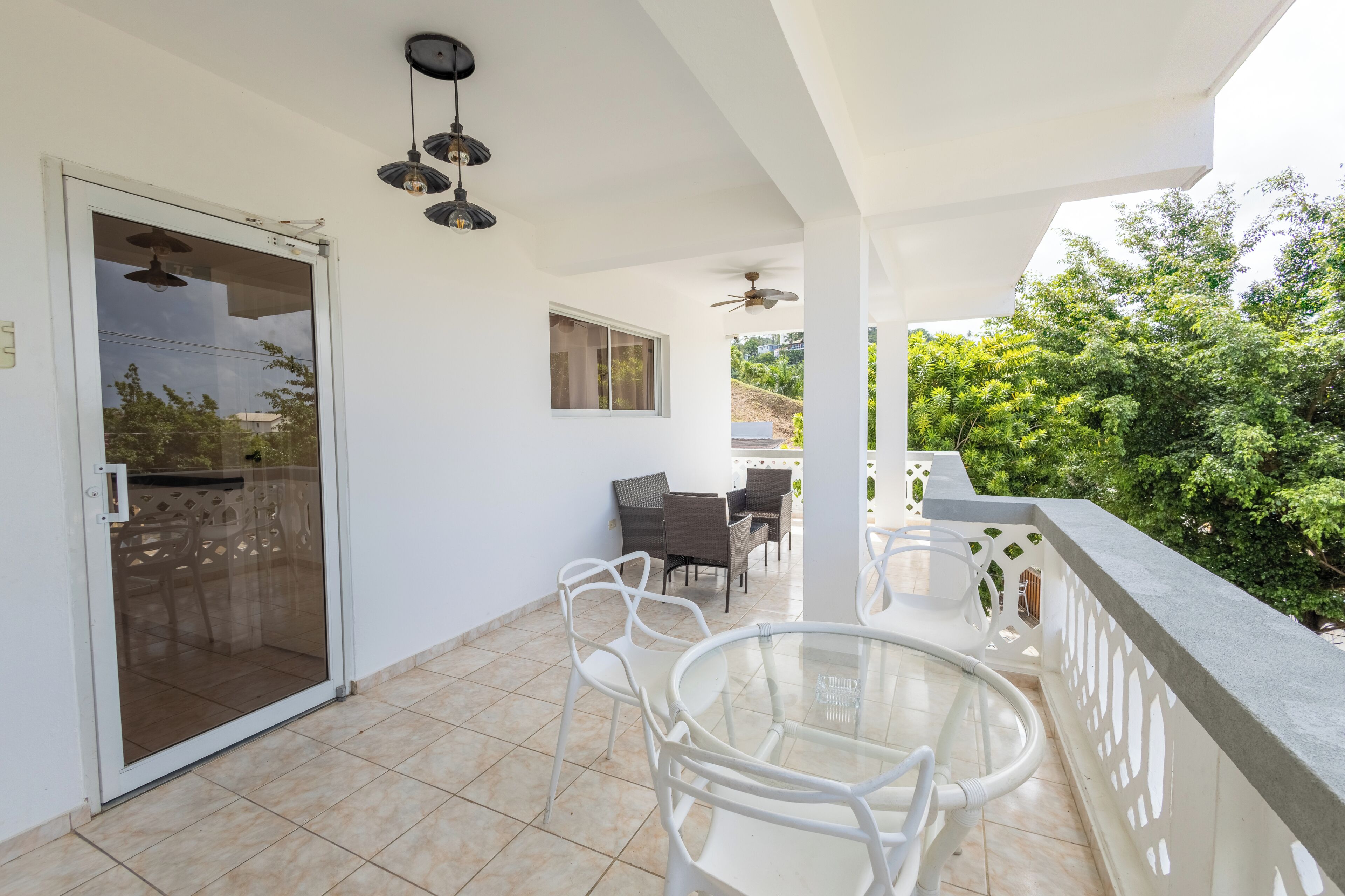 A balcony with a glass door, white chairs, a glass table, and a ceiling fan.