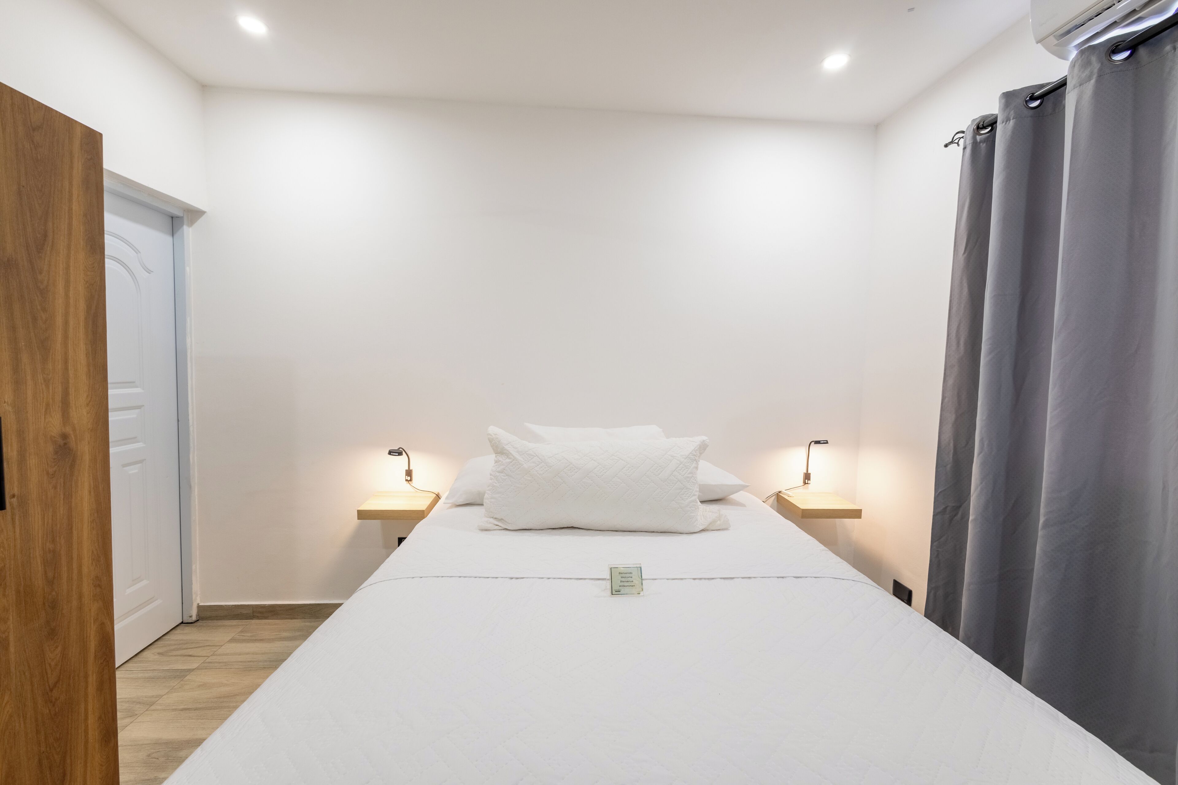 Uma cama de solteiro com colchão e almofadas brancas, com duas mesas de cabeceira com luminárias, em um quarto com porta de madeira e cortina cinza.