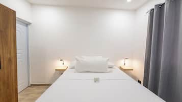 Uma cama de solteiro com colchão e almofadas brancas, com duas mesas de cabeceira com luminárias, em um quarto com porta de madeira e cortina cinza.