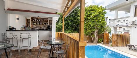 Zona junto a la piscina con bar, asientos y vista a un árbol.