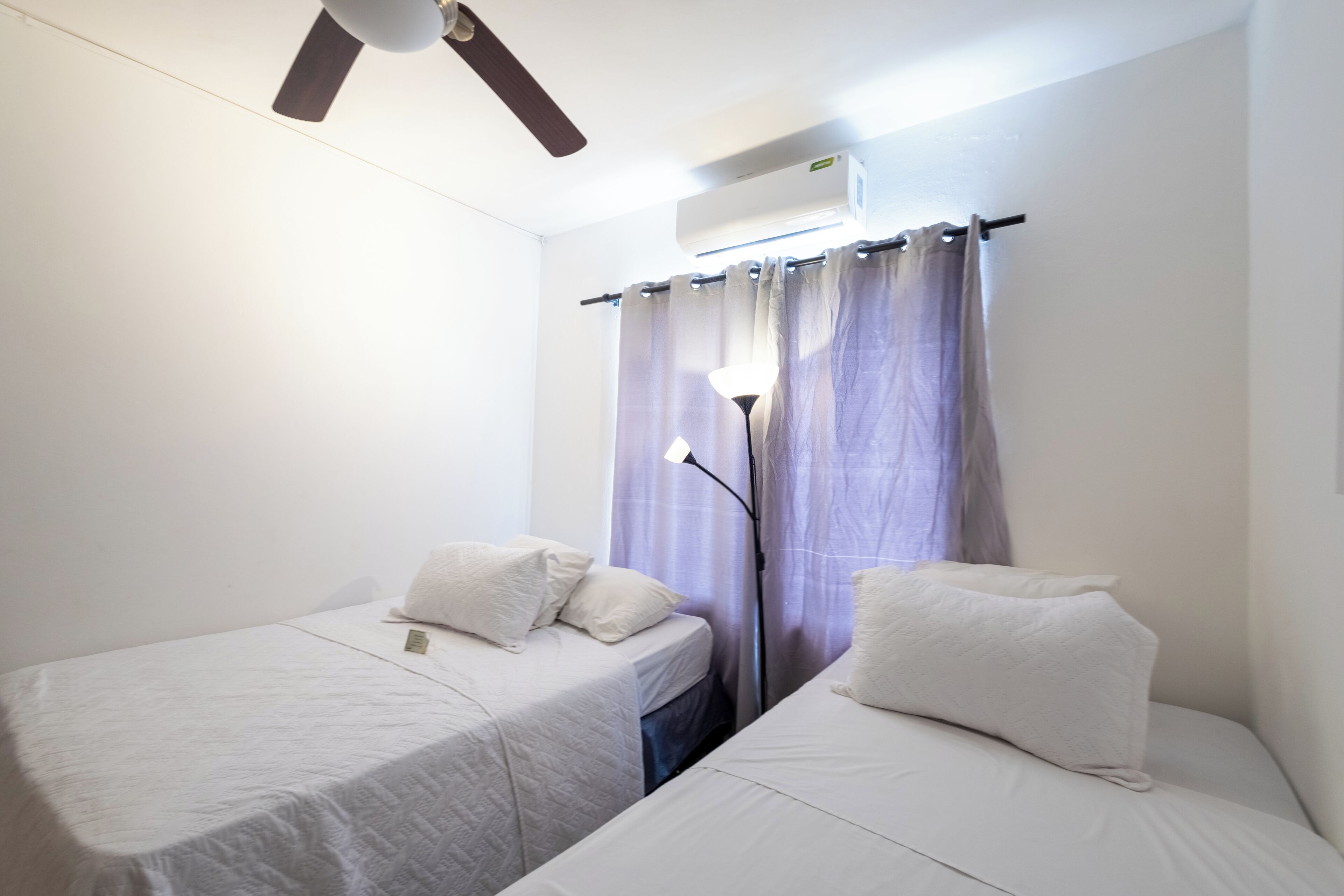 A room with two single beds, a ceiling fan, an air conditioning unit, and a lamp.