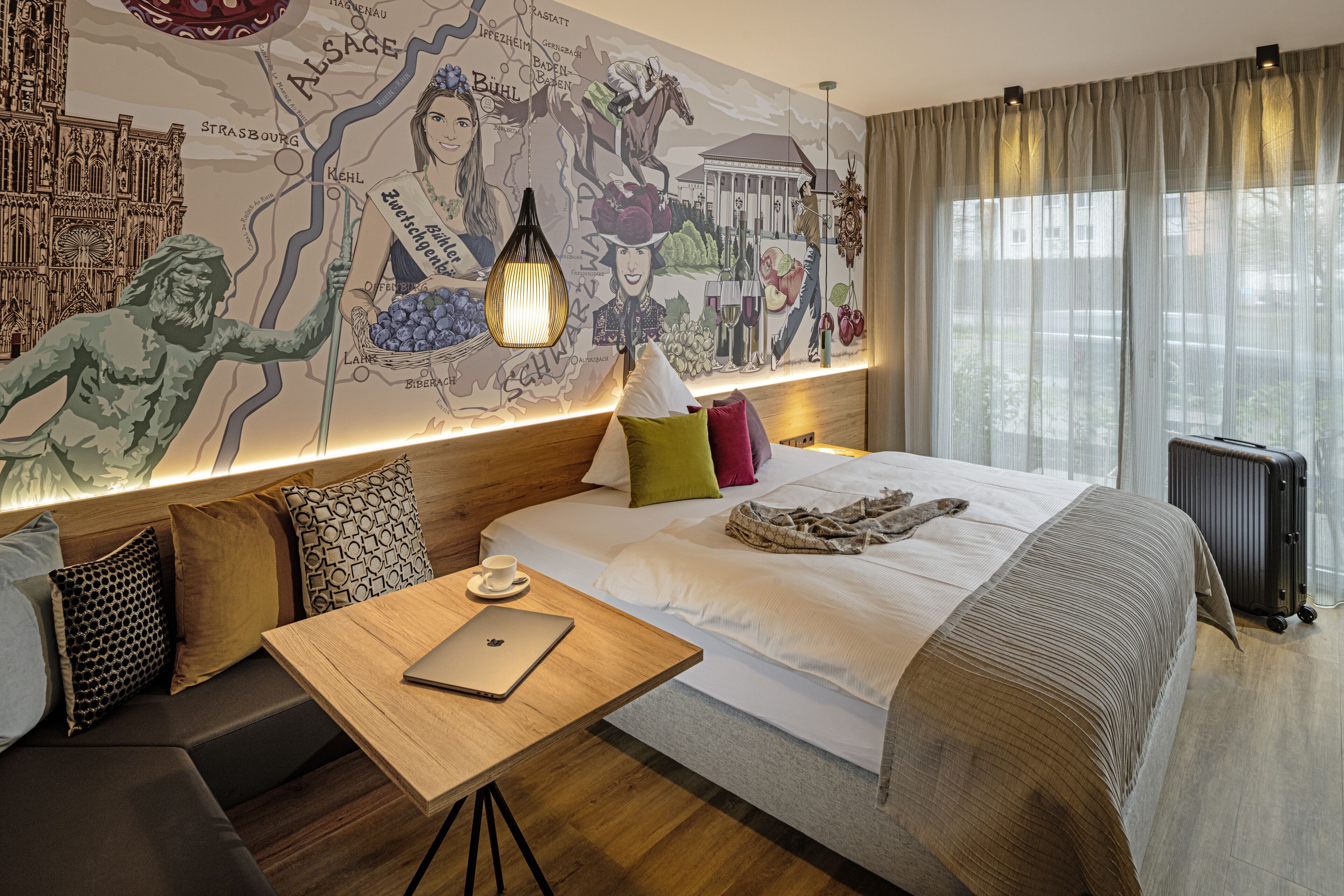Ein Hotelzimmer mit einer großen Wandmalerei, einem hölzernen Kopfteil, einem Bett mit vielen Kissen, einem Tisch mit einem Laptop und einem Heizkörper.