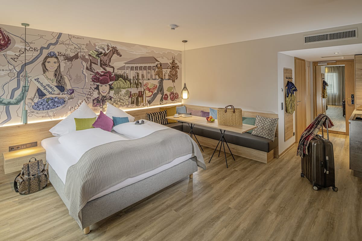 Una habitación de hotel moderna con una cama grande, un área de descanso con una mesa, un mural de un mapa en la pared y una maleta en el suelo.
