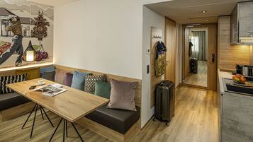 Una habitación de hotel moderna con una mesa de comedor de madera, un sofá con cojines de colores, un perchero, una maleta y una zona de cocina con un frutero.