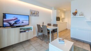 Un soggiorno moderno con una TV a schermo piatto, un'area pranzo con tavolo e sedie, e una cucina con mobili bianchi e forno a incasso.