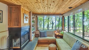 Interior acogedor de una cabaña con chimenea, techo de madera, ventanas amplias y una zona de estar con cojines.
