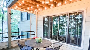Un patio al aire libre con una mesa redonda y sillas, luces colgantes y grandes ventanales.