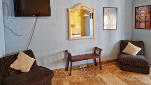 A room with a flat-screen TV, a large mirror, two patterned sofas, a bench, and a wall-mounted air conditioner. - Apartamento El Duque (Córdoba)
