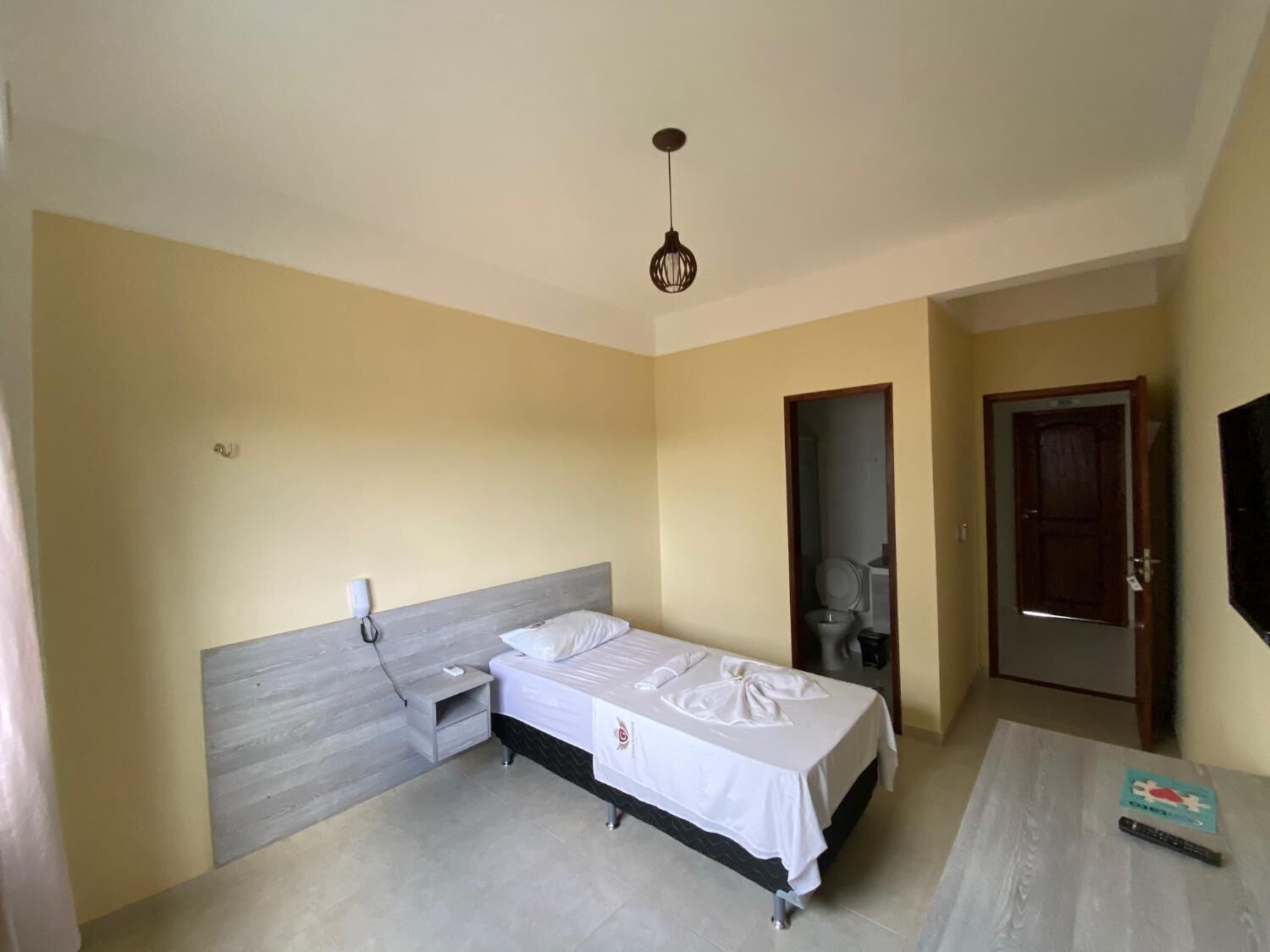 A small, clean room with a single bed, a bedside table, a nightstand, a wall-mounted TV, and a door leading to another room.