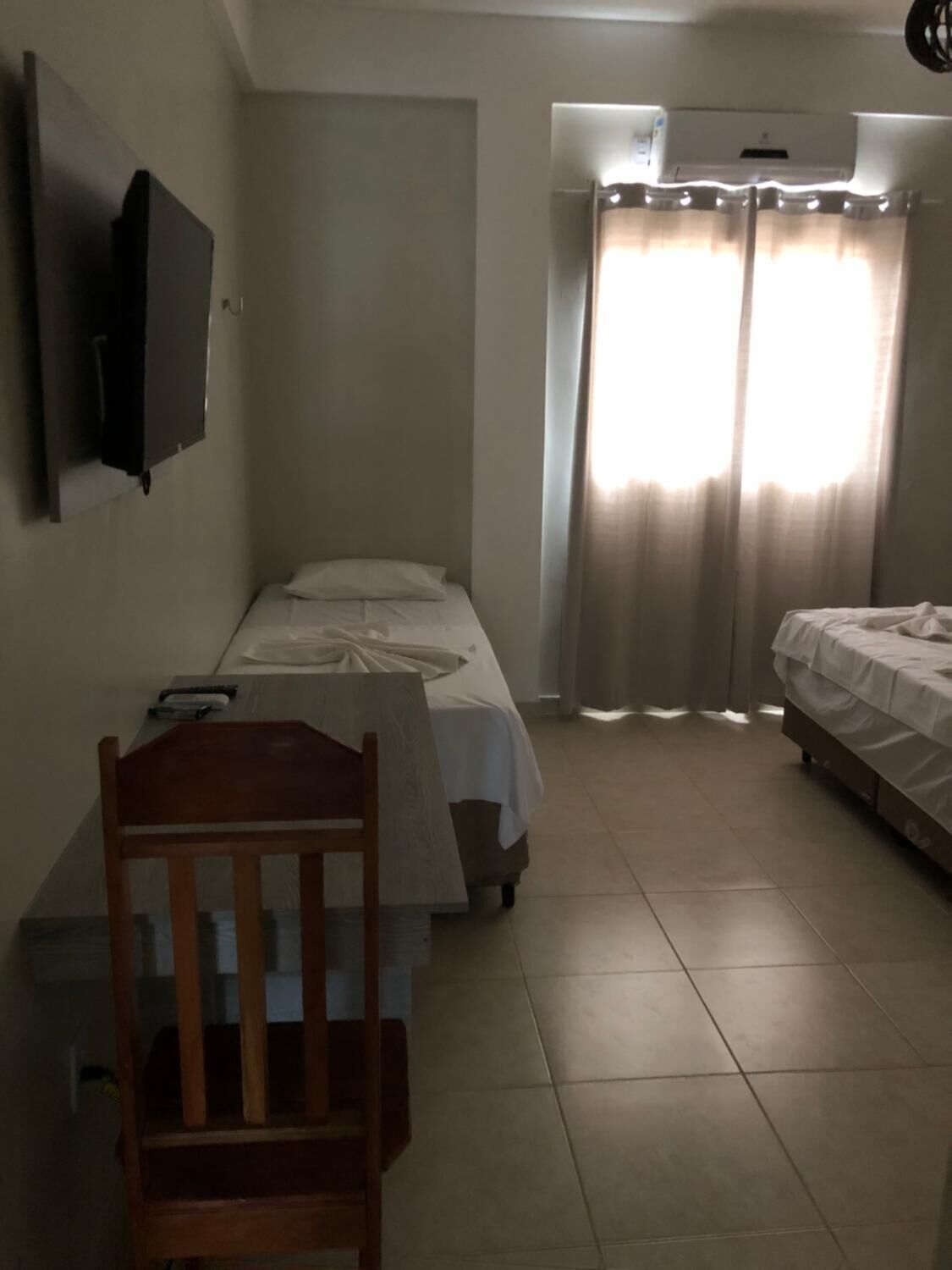 A small, well-lit room with two beds, a wooden chair, a flat-screen TV, and a window with curtains.