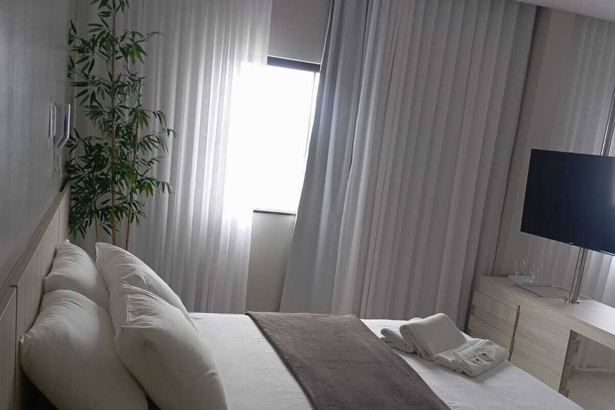 A hotel room with a large bed, a bedside table with a remote control, a flat-screen TV, and a potted plant.