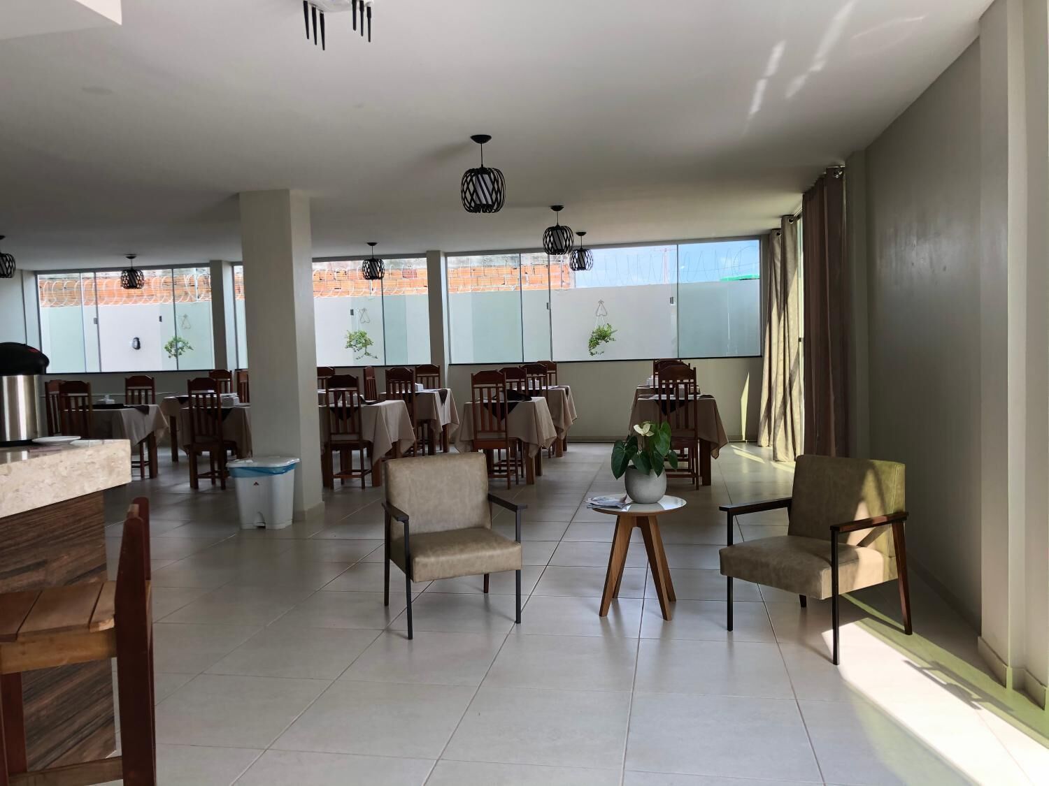 A spacious dining area with multiple tables and chairs, large windows with views of a brick building, and hanging pendant lights.
