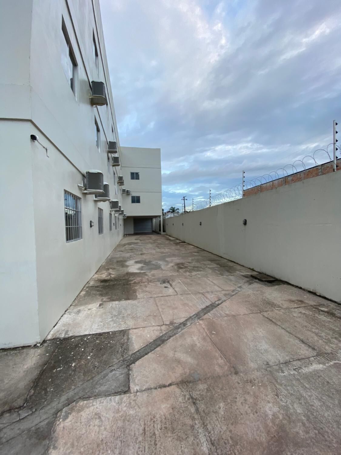 Un pasillo con piso de baldosas, un edificio blanco con unidades de aire acondicionado y una valla de alambre de púas.