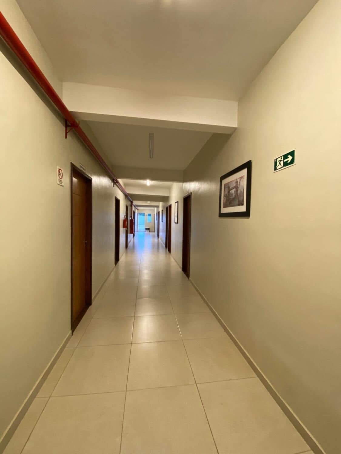 A long, narrow hallway with doors on both sides, a tiled floor, and a green emergency exit sign on the right wall.