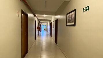 A long, narrow hallway with doors on both sides, a tiled floor, and a green emergency exit sign on the right wall.