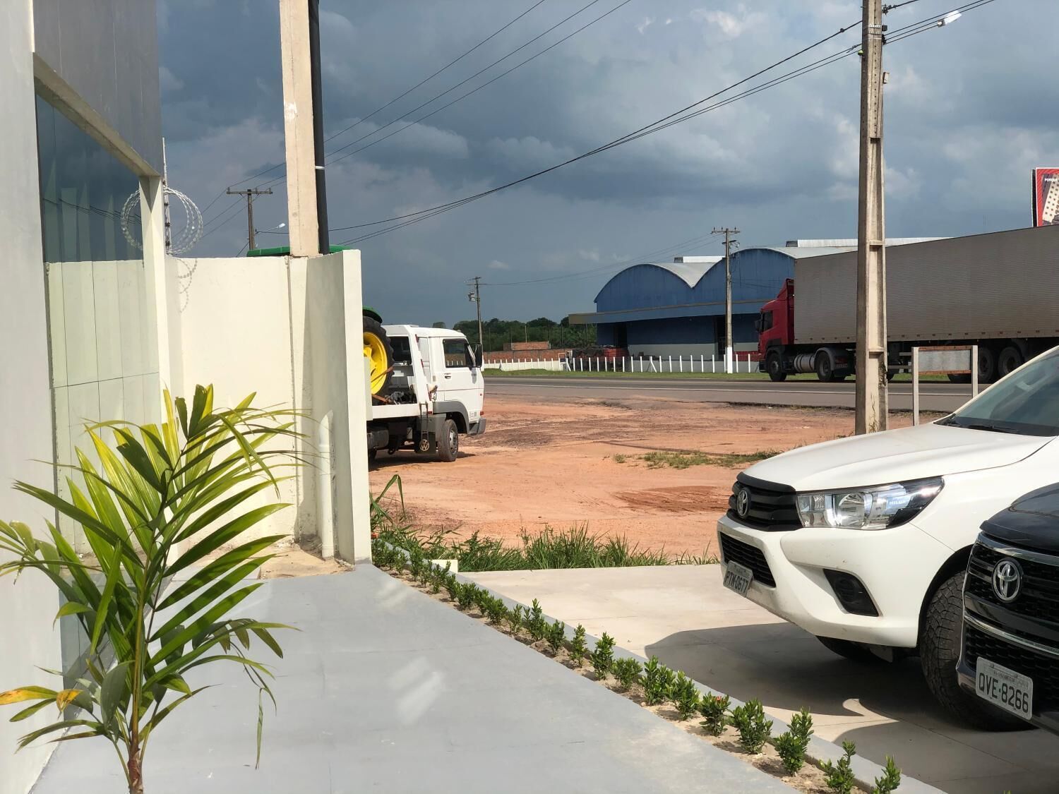 A parking area with a white car and a white truck, a building with a blue roof, and a plant in the foreground.