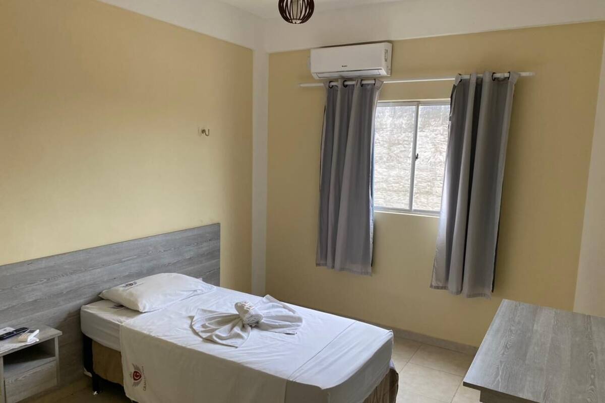 A hotel room with a bed, a desk, an air conditioning unit, and a window with curtains.