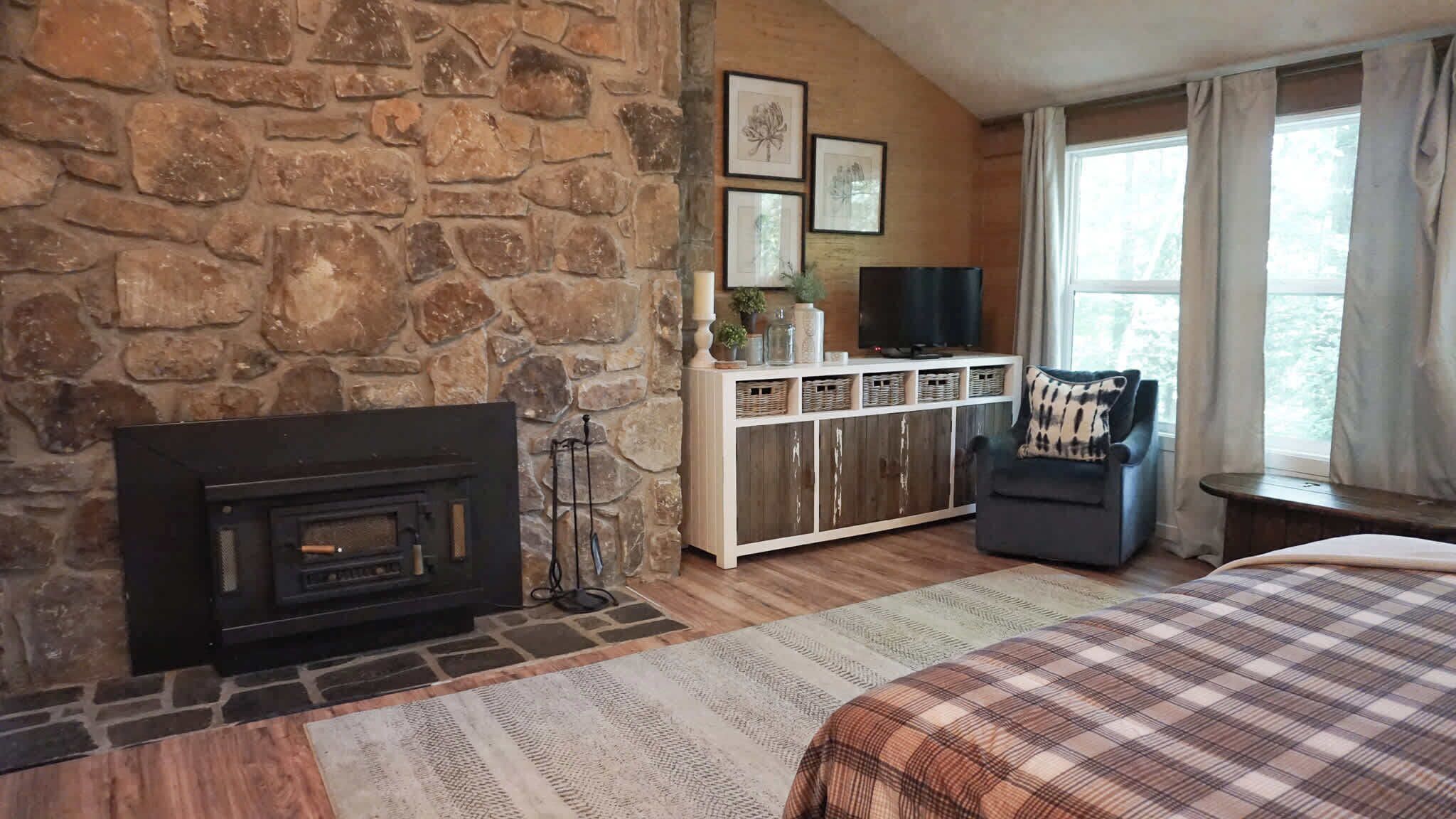 A cozy room with a stone fireplace, a comfortable armchair, and a bed with a plaid bedspread.