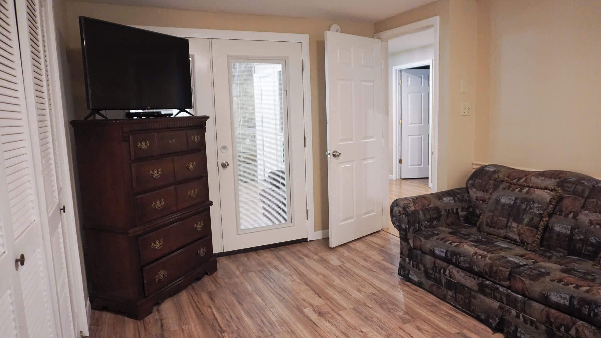 A room with a wooden dresser, a television on top, a patterned sofa, and a door leading to another room.