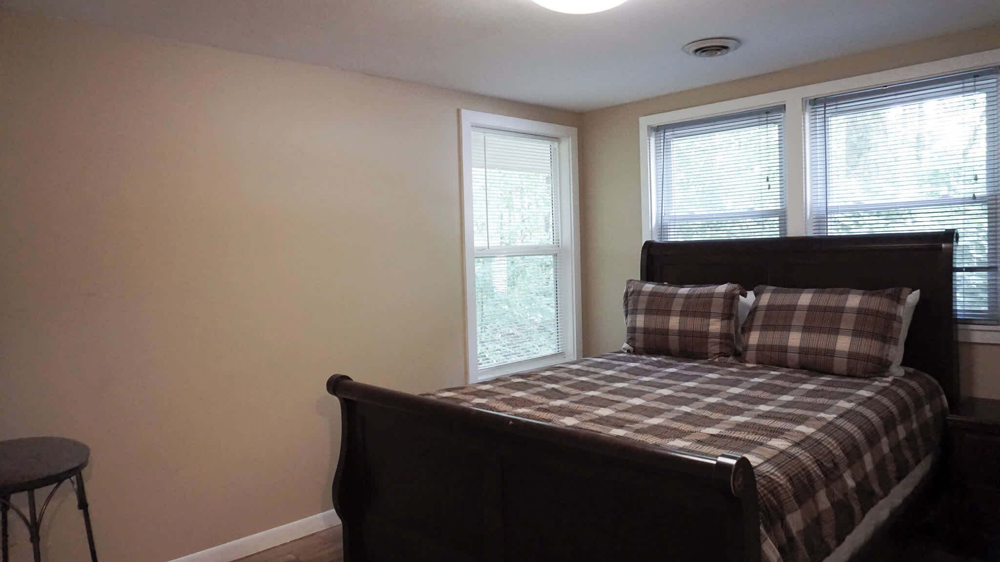 A bedroom with a bed, a chair, and two windows with blinds.