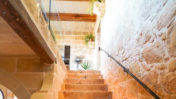 A stone staircase with a metal handrail, leading to a room with a stone wall and a fan in the background.