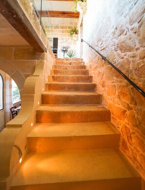 Un escalier en pierre avec une rampe métallique, menant à une pièce dont un mur est en pierre et où l’on aperçoit un ventilateur au fond.