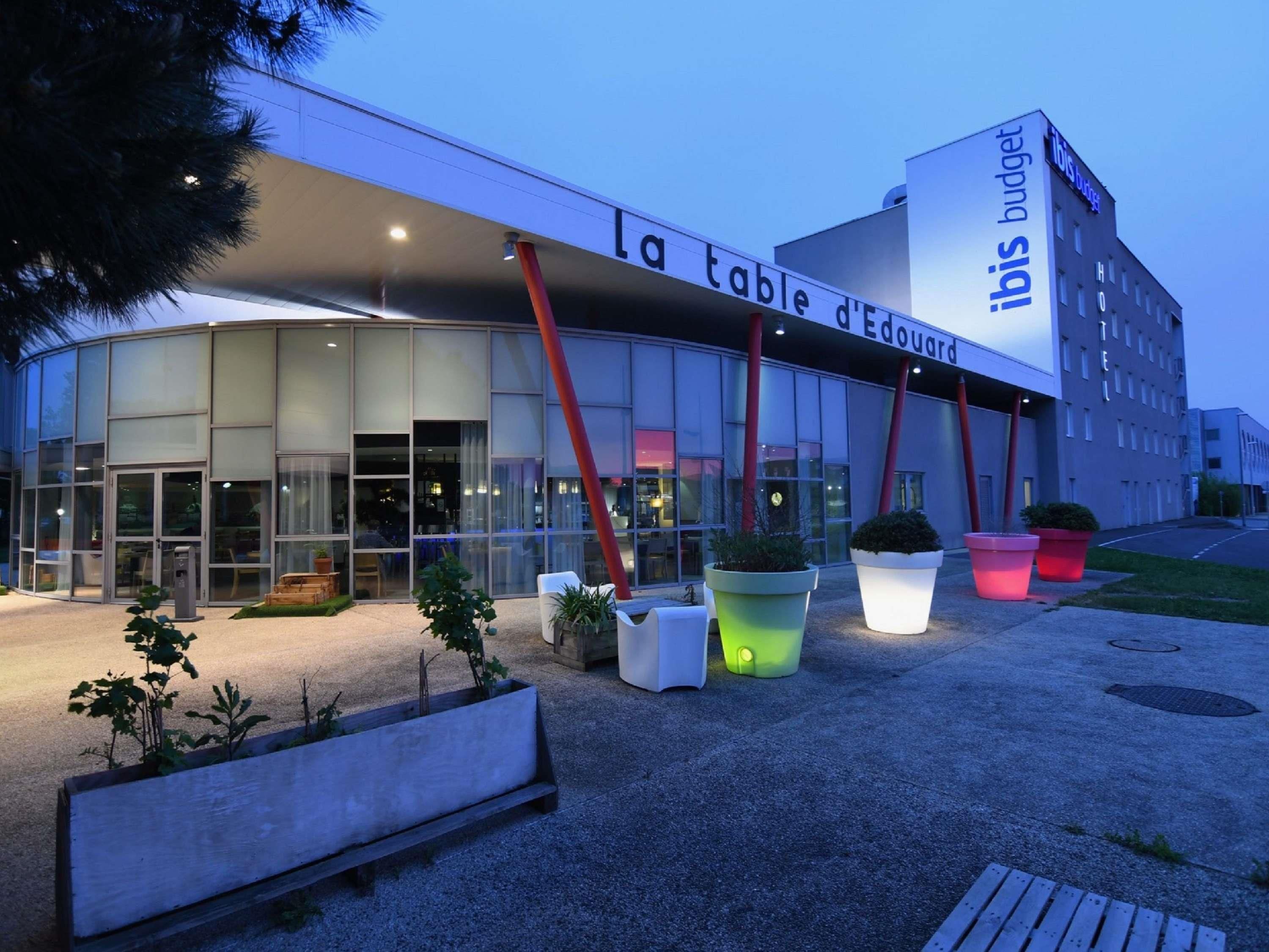 Un moderno edificio de hotel con fachada de vidrio, una entrada destacada y zonas de estar al aire libre con jardineras.