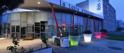 Un moderno edificio de hotel con fachada de vidrio, una entrada destacada y zonas de estar al aire libre con jardineras.