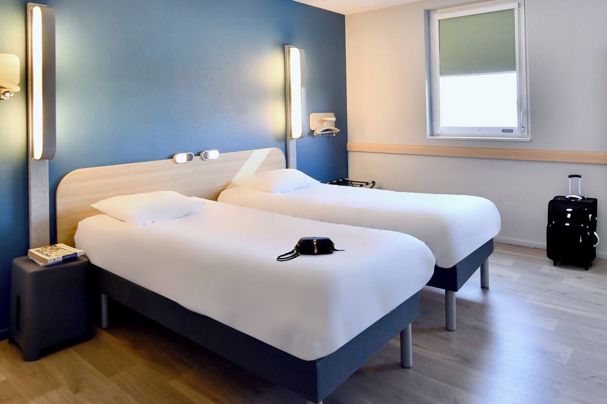 A hotel room with two beds, a nightstand, a suitcase, and a window with blinds.