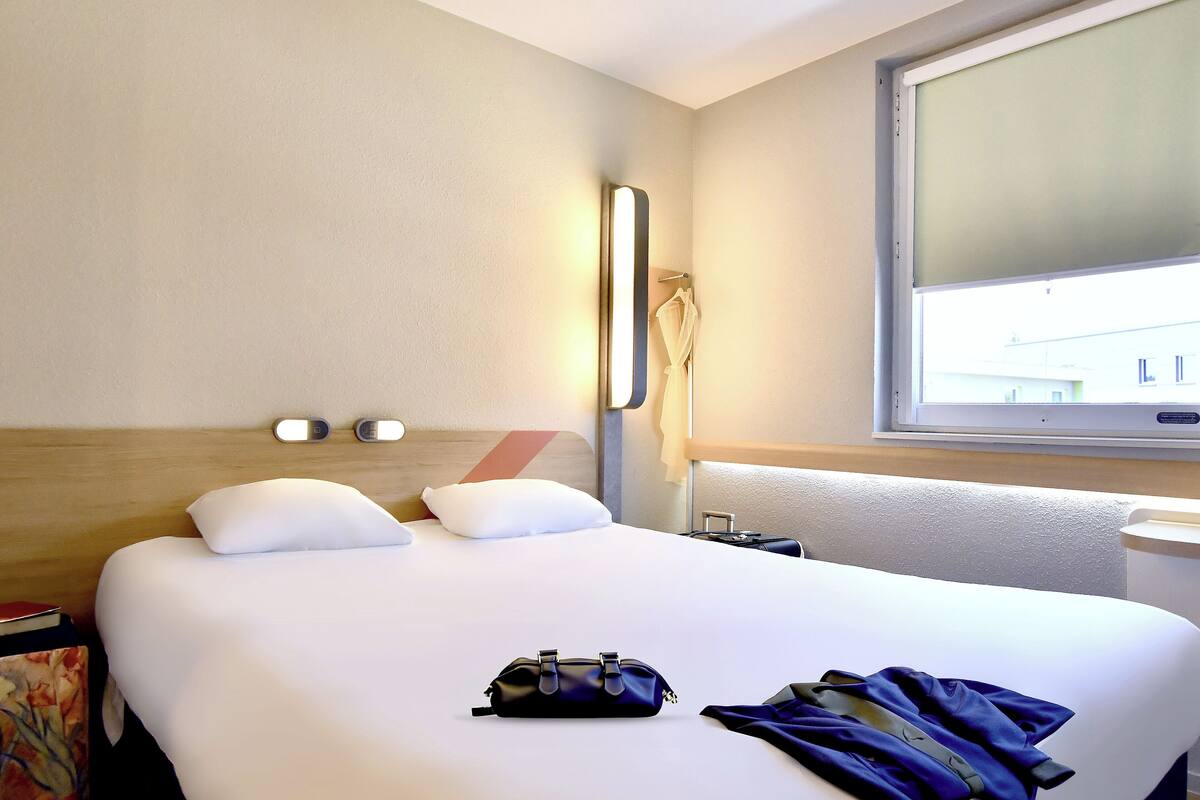 A hotel room with a large bed, a bedside table, a window, and a wall-mounted light fixture.