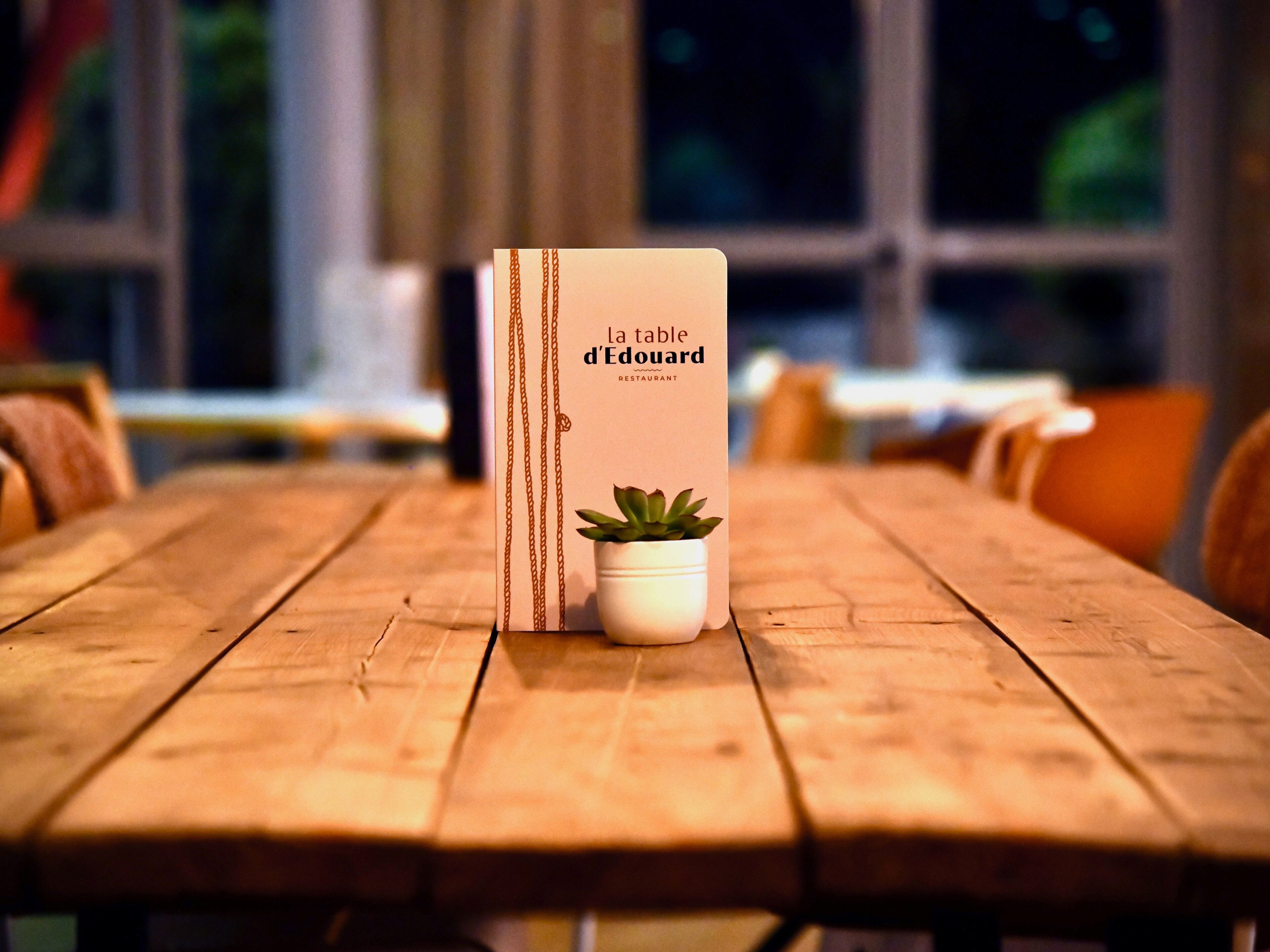 Una mesa de madera con un atril de menú que muestra el nombre del restaurante 'la table d'Edouard' y una pequeña planta en maceta.
