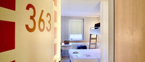 Una habitación de hotel con literas, una mesita de noche con lámpara y una ventana.