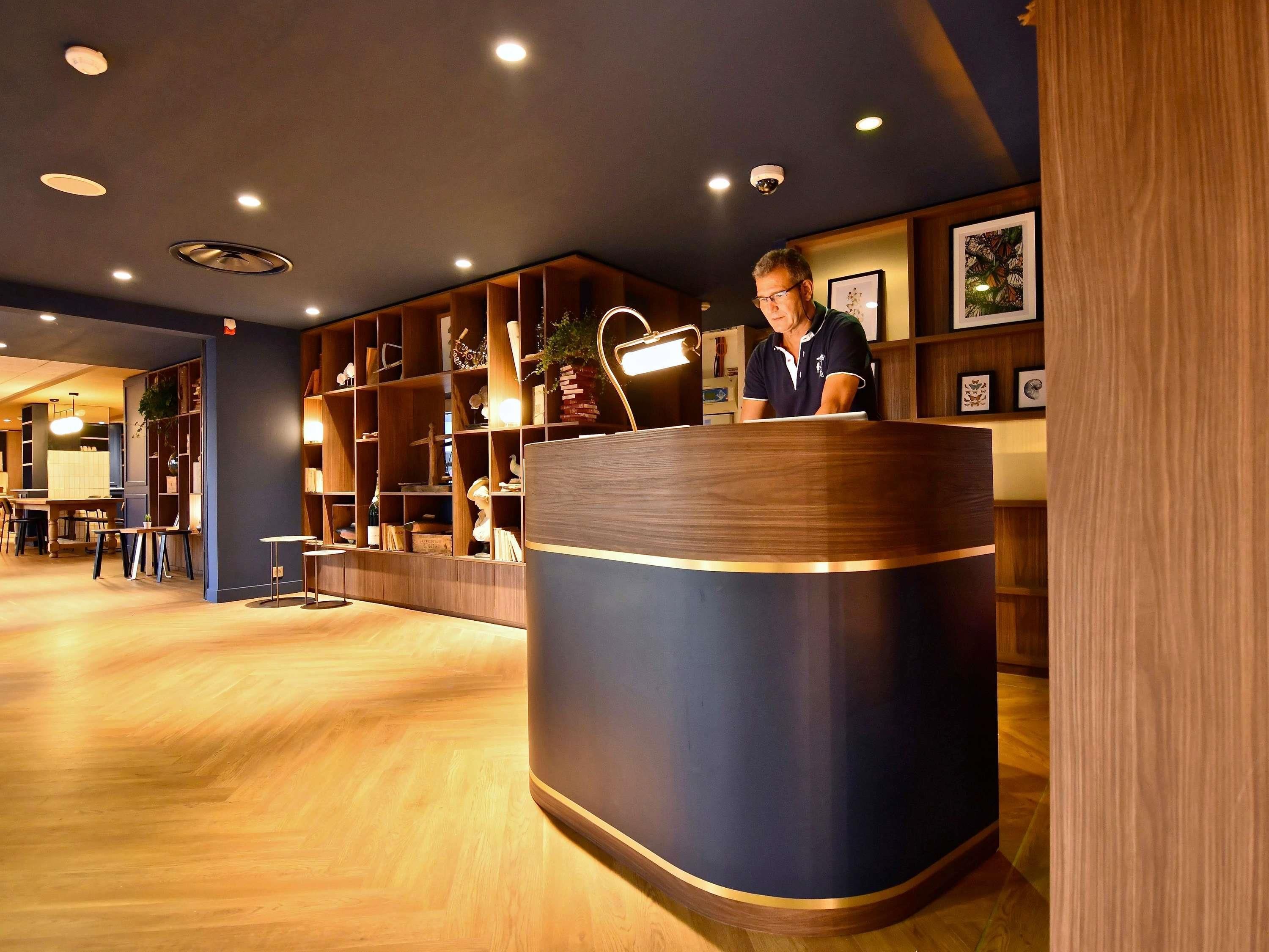A reception area with a wooden reception desk, shelves with various items, and a seating area with tables and chairs.