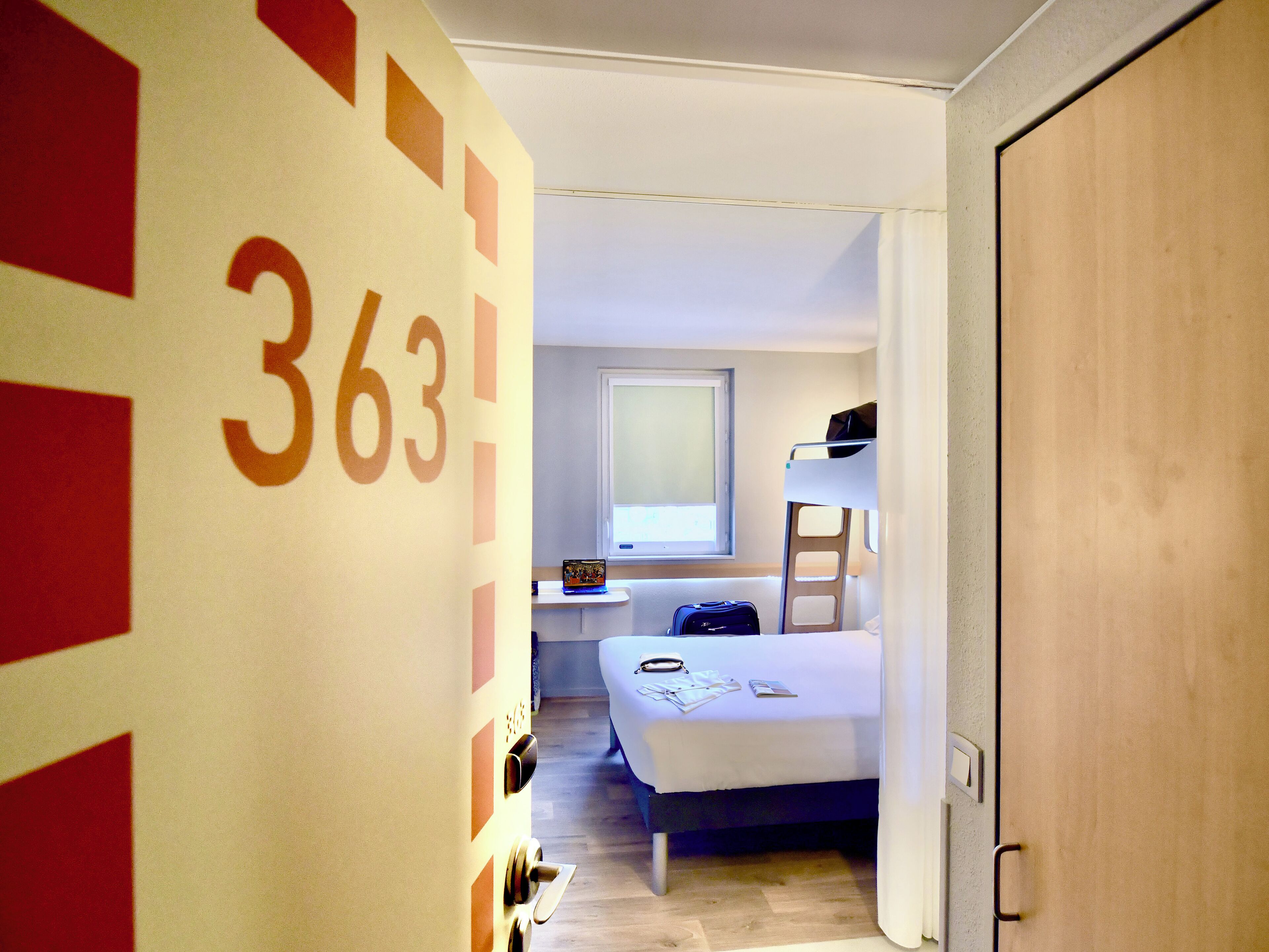 A hotel room with a bunk bed, a bedside table with a lamp, a window, and a door with the number 363.
