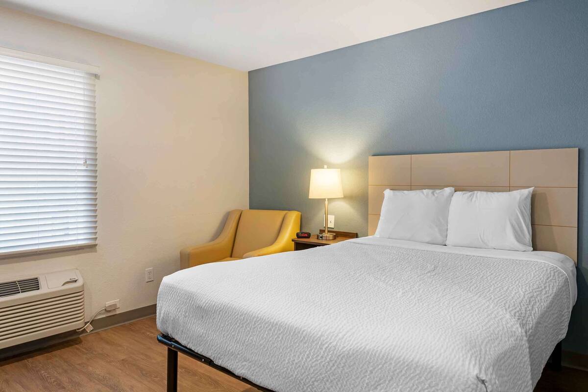 A hotel room with a bed, a chair, a nightstand with a lamp, and a window with blinds.